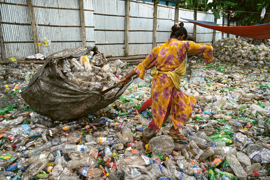Schauplatz Asien, eine Person steht auf einem Berg aus Plastik und versucht eine große Tasche voller Plastik wegzuziehen.