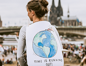 Vorschaubild: Junge Frau mit Schild auf dem Rücken, Aufschrift: "Time is running out". Im Hintergrund der Kölner Dom
