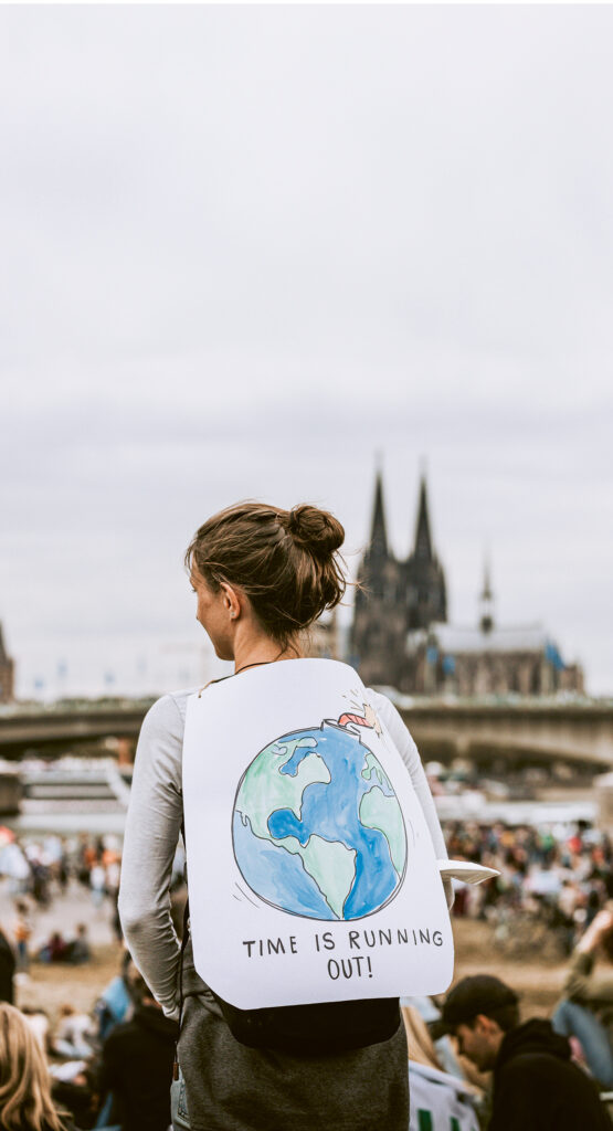 Junge Frau mit Schild auf dem Rücken, Aufschrift: "Time is running out". Im Hintergrund derKölner Dom